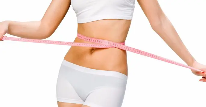 A woman in a white sports bra and underwear measuring her waist with a pink measuring tape.