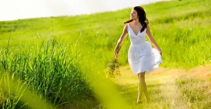 A person in a white dress walks through a lush green field with long grasses, holding a small bouquet of yellow flowers. The environment is bathed in bright sunlight, and the person has dark hair and a playful pose, suggesting movement through the natural setting.