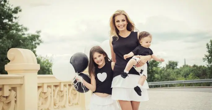 A family wearing coordinated black and white outfits, with two children holding balloons, standing on a bridge with ornate railings, under a cloudy sky.