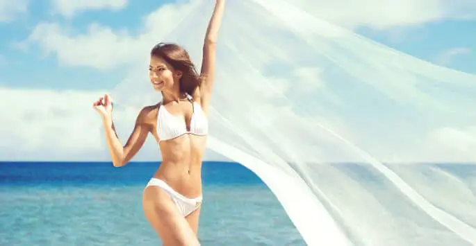 A person wearing a white bikini standing on a beach with arms raised holding a flowing white fabric against a backdrop of the blue sea and sky.