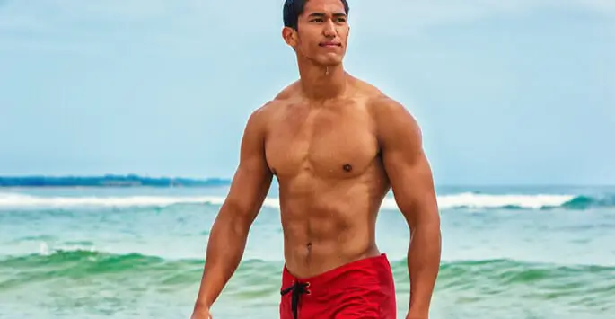 A shirtless man with a fit physique wearing red swim shorts standing on a beach with the ocean in the background.