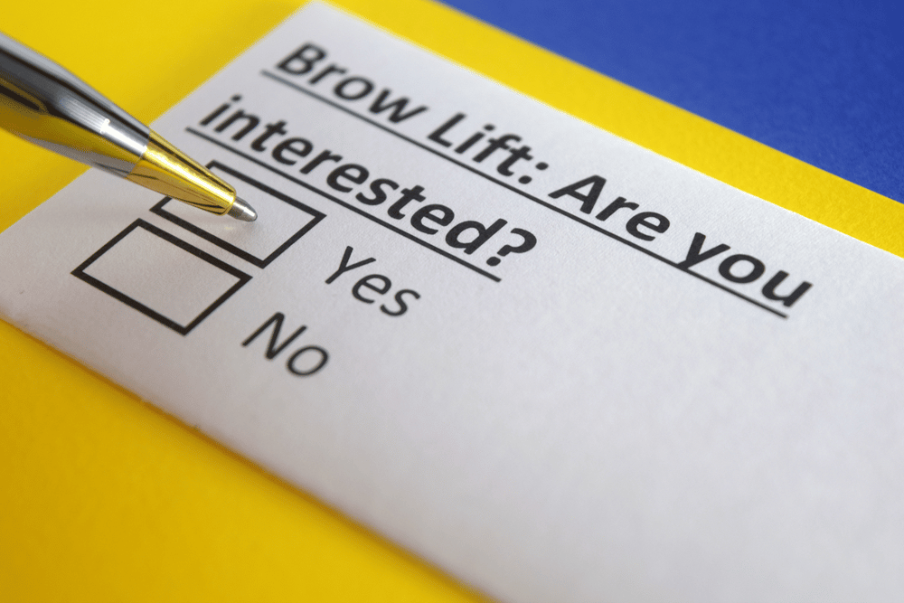 A close-up of a paper with the text "Brow Lift: Are you interested?" with checkboxes for "Yes" and "No", and a pen positioned over the "Yes" checkbox.