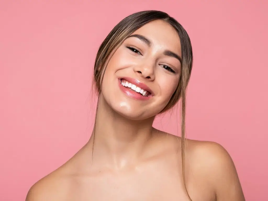 A person with bare shoulders against a pink background with a ponytail hairstyle visible from the side.