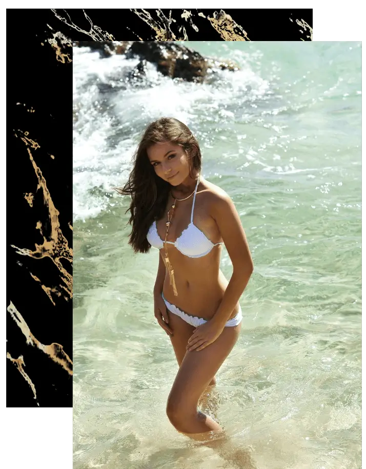A woman in a white bikini stands in shallow ocean water, smiling, with waves splashing around her.